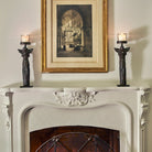Two wax cast iron double angel on stand with mouth blown glass insert with lit candles on top of a white chimney. A photo in the center.