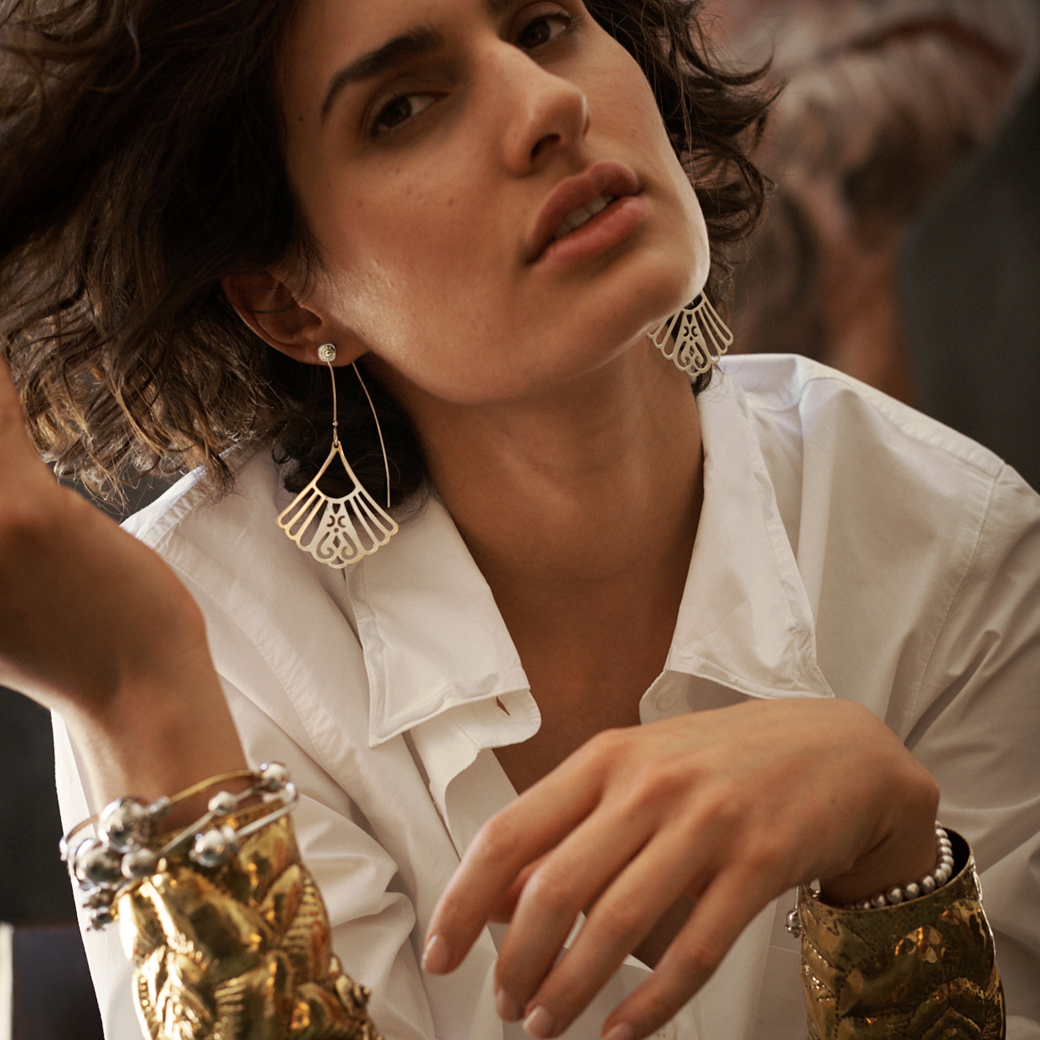 A woman playing with her hair.looking aloof. Her hand is in the lower center of the photo. She is wearing a white shirt and two silver earrings with a fan shape and a hook.