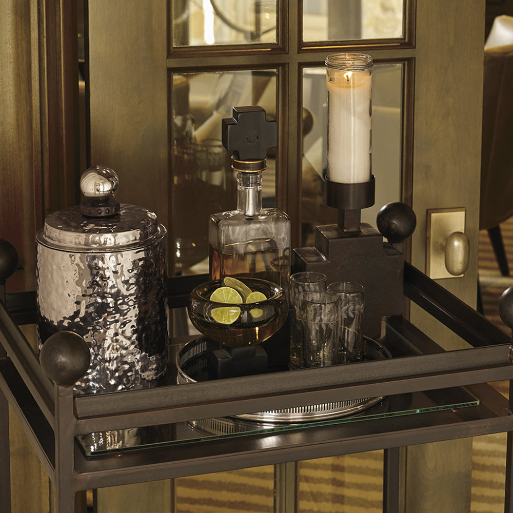 A tray with a hammered  steel ice bucket with lid, a decanter with an iron cross topper, a  candle on top of an iron cross, a bowl with sliced lime and three glasses. On the background is a door.