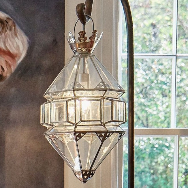 A aluminum and glass lantern hanging from a iron hook. On the background is a window.